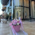 Petite Петит корзина с нежными цветами перед бутиком Chanel на солнечной набережной.